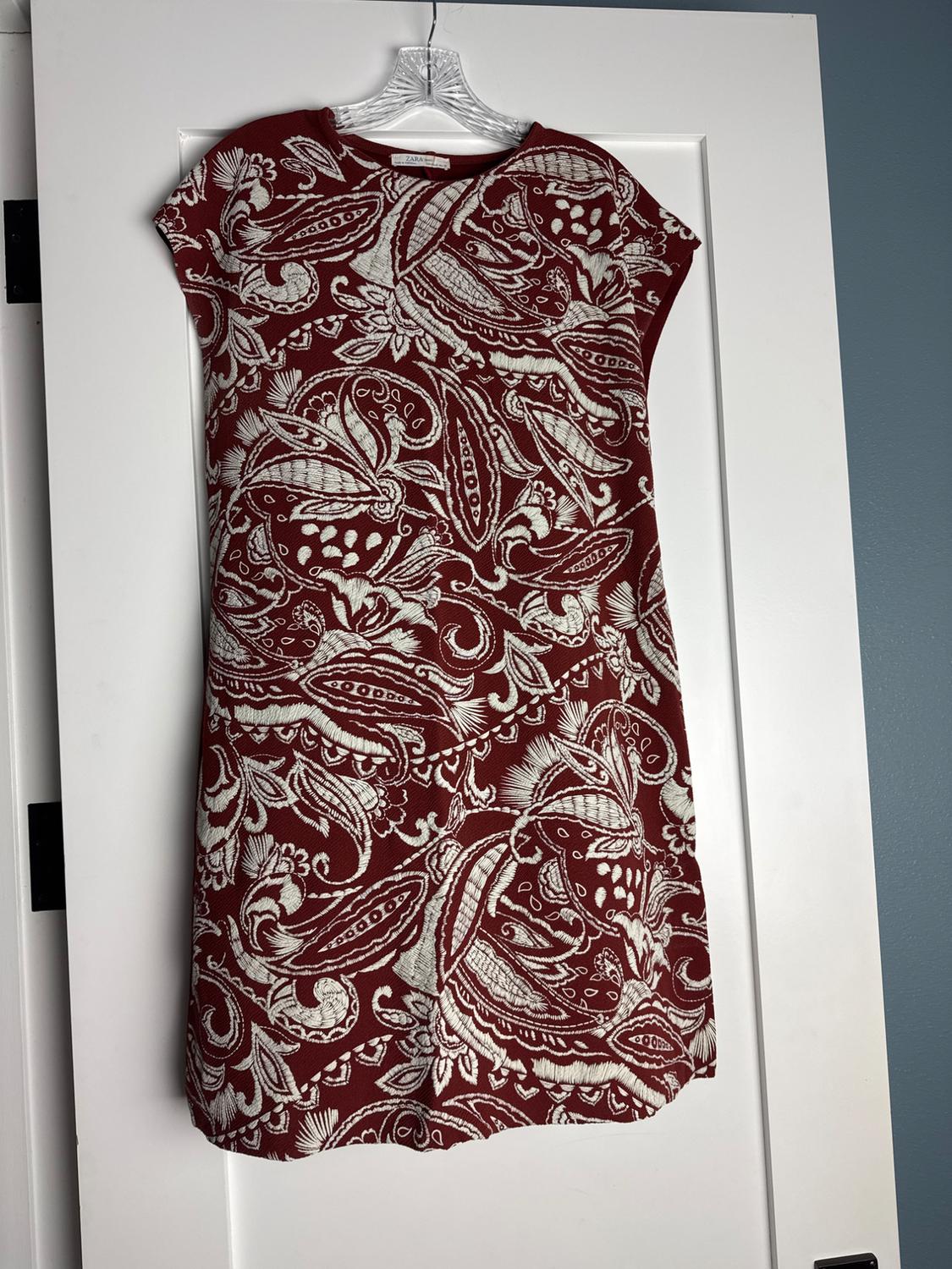 Maroon sleeveless short dress with a white print hanging on a hanger over a white door. Label with the text ZARA.