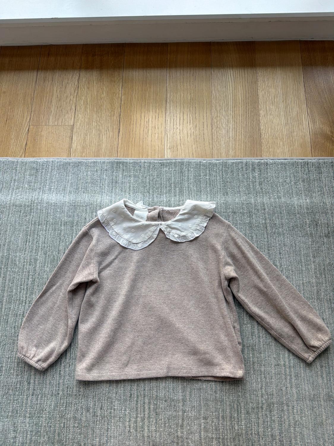 Beige long-sleeved jumper with a white ruffled collar, placed on a grey rug and light wooden flooring.