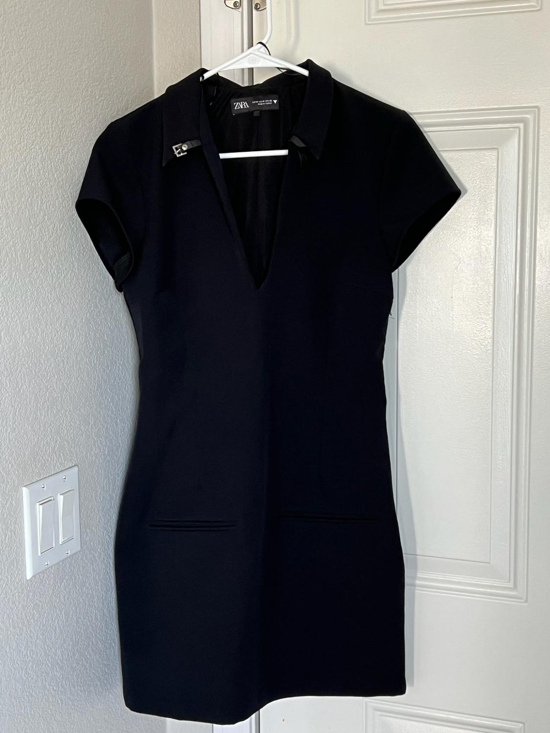 Black short-sleeved dress, V-neck, buckle collar, and pockets, hanging on a white door. Tag with ZARA text.