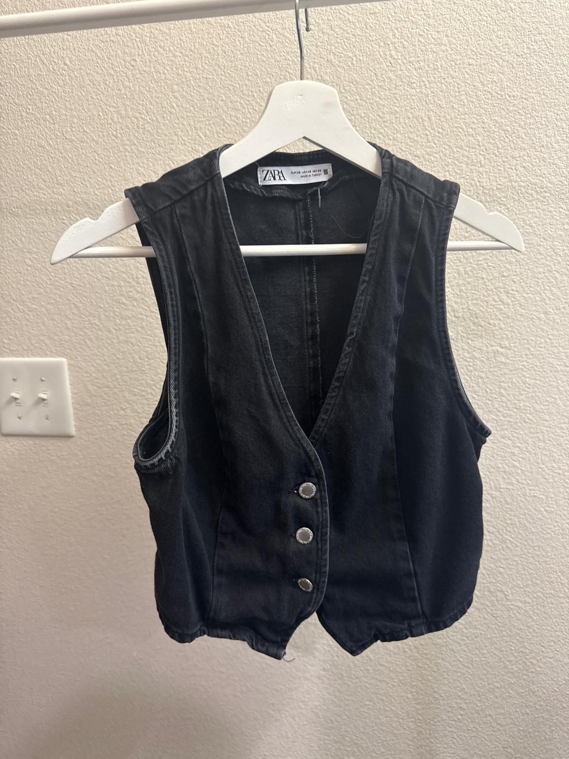 Black denim waistcoat with silver buttons hanging on a white hanger, against a light-coloured wall. Visible label: ZARA.