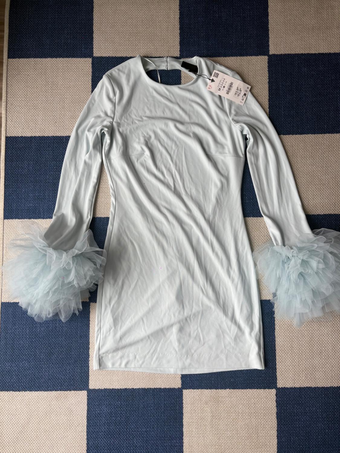 Light blue dress with long sleeves and tulle cuffs, on a blue and beige checkered rug. Visible tag on the collar.