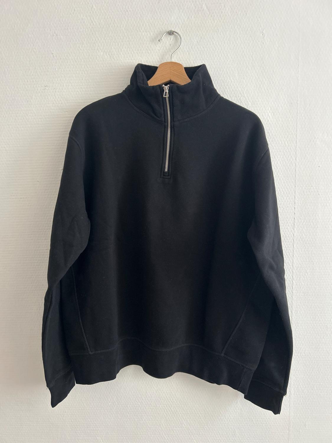 Black sweatshirt with a zip-up neck, hanging on a wooden hanger against a white wall.