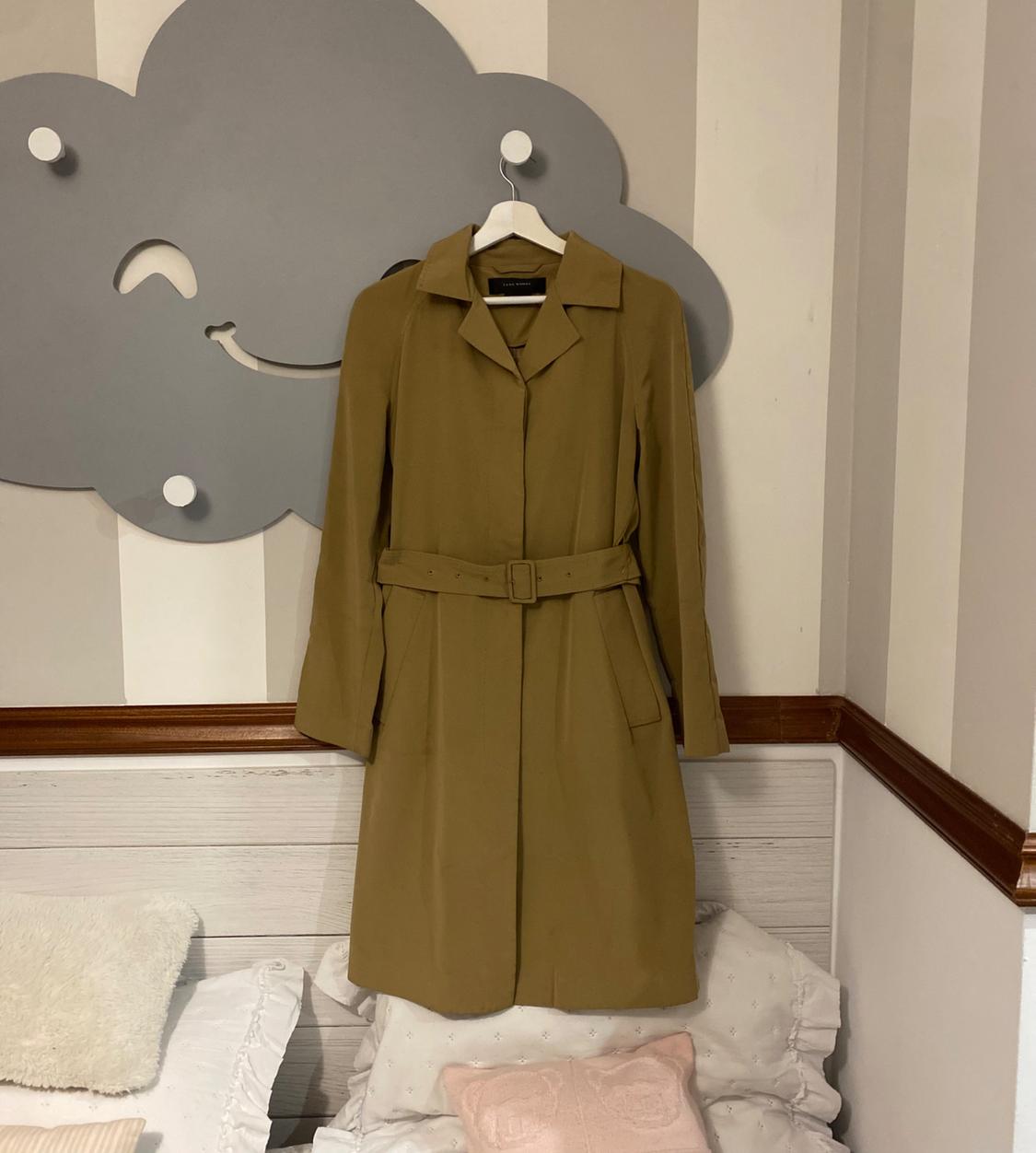 Light brown coat with belt hanging on a white hanger against a wall with grey cloud decor and bed with cushions.