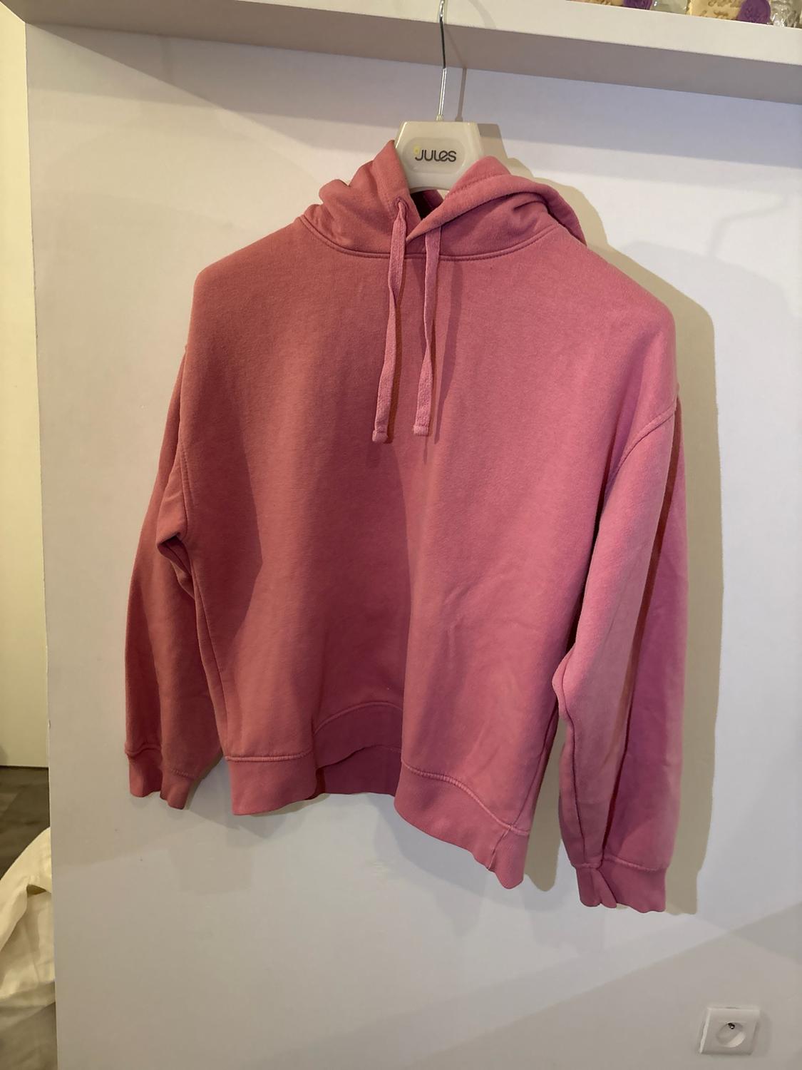 Pink hoodie hanging on a white hanger with the text JULES, against a white wall.