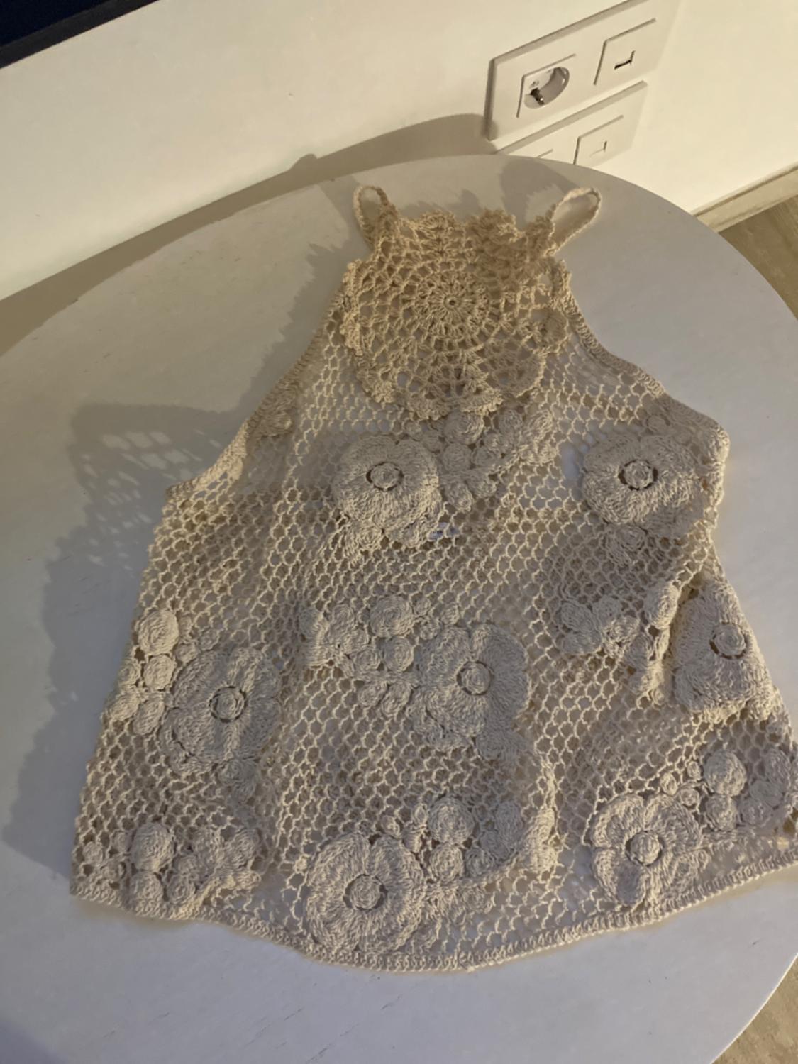 Beige crochet top with floral design on a white table. Soft light creates shadows on the wall and surface. Plug sockets visible in the background.