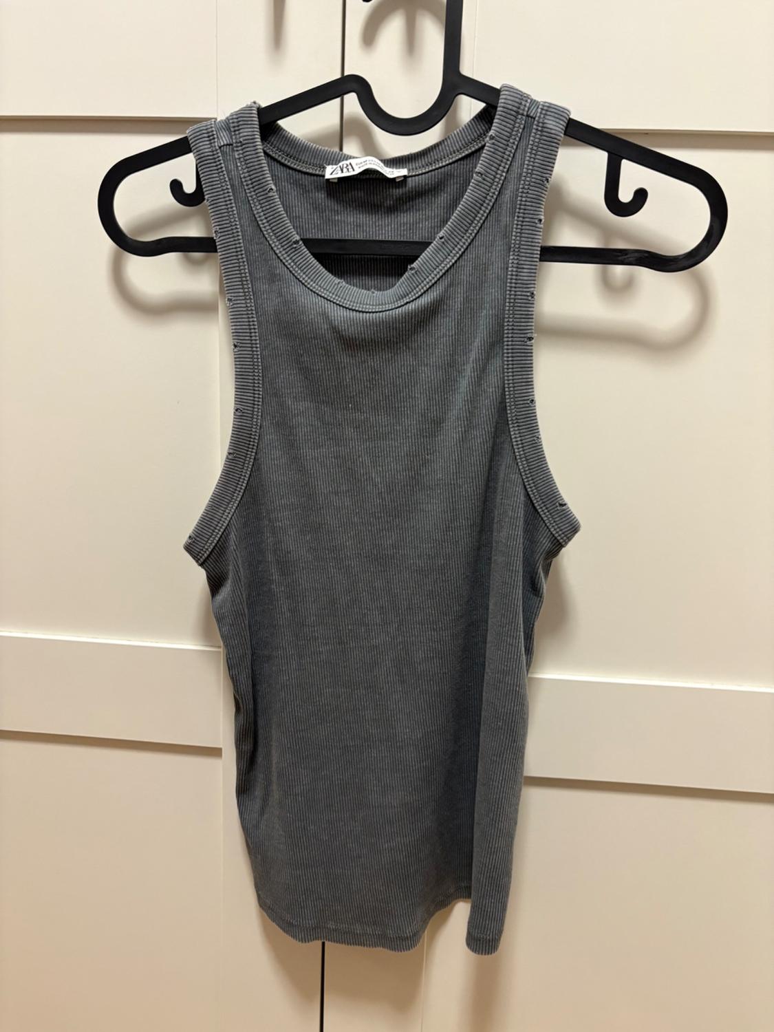 Grey ribbed vest top with distressed edges on a black hanger in front of a white wardrobe. Text: ZARA.