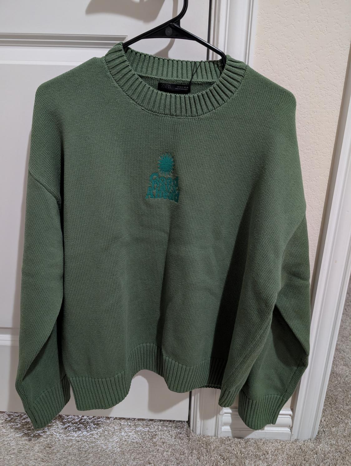 Green jumper hanging on a black hanger, with an embroidered design on the chest that says "Good Vibes Always". White door background.