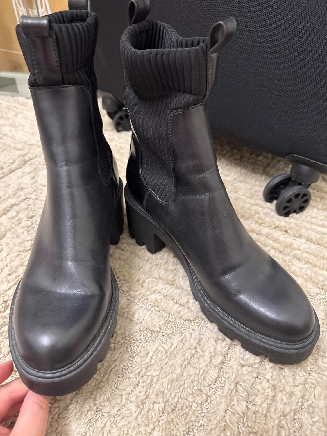 Black leather boots with a chunky sole and ribbed elastic, on a beige carpet. Background with a black wheeled suitcase.