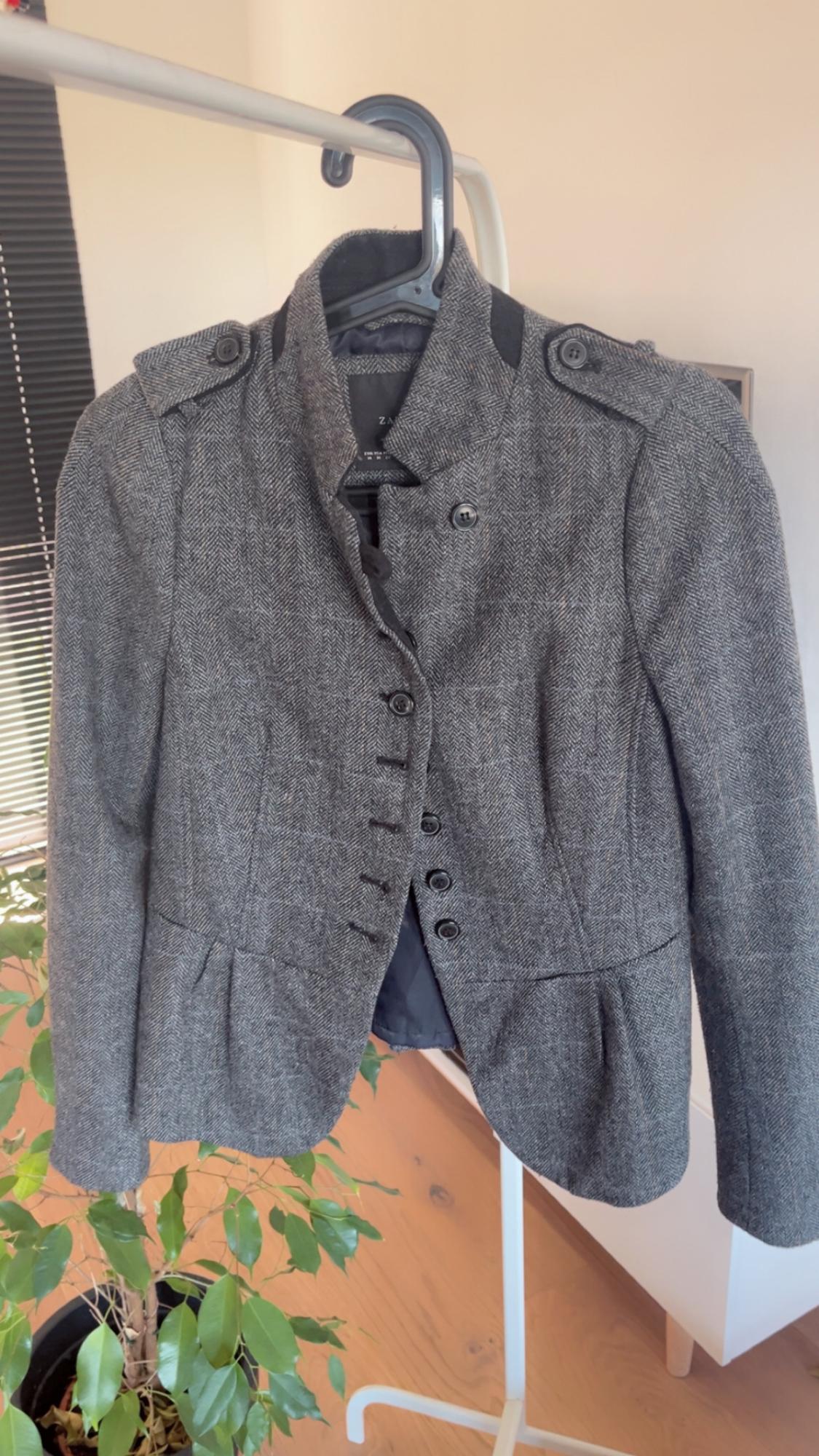 Grey button-up jacket hanging on a coat rack, with a green plant and black blind in the background. Visible tag: ZARA.