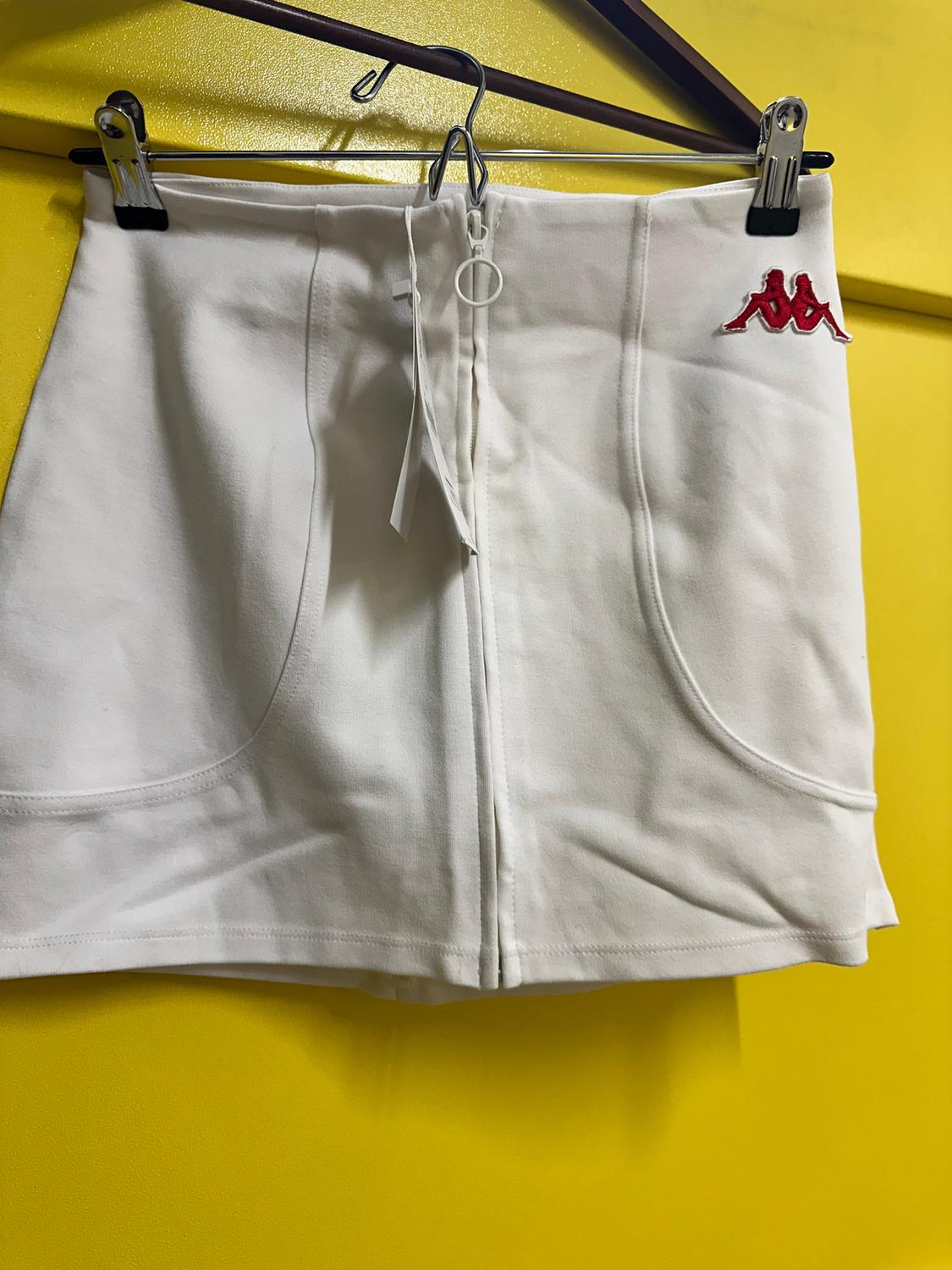 White mini skirt with a front zip and a red logo on the waistband, hanging on a hanger against a yellow background.
