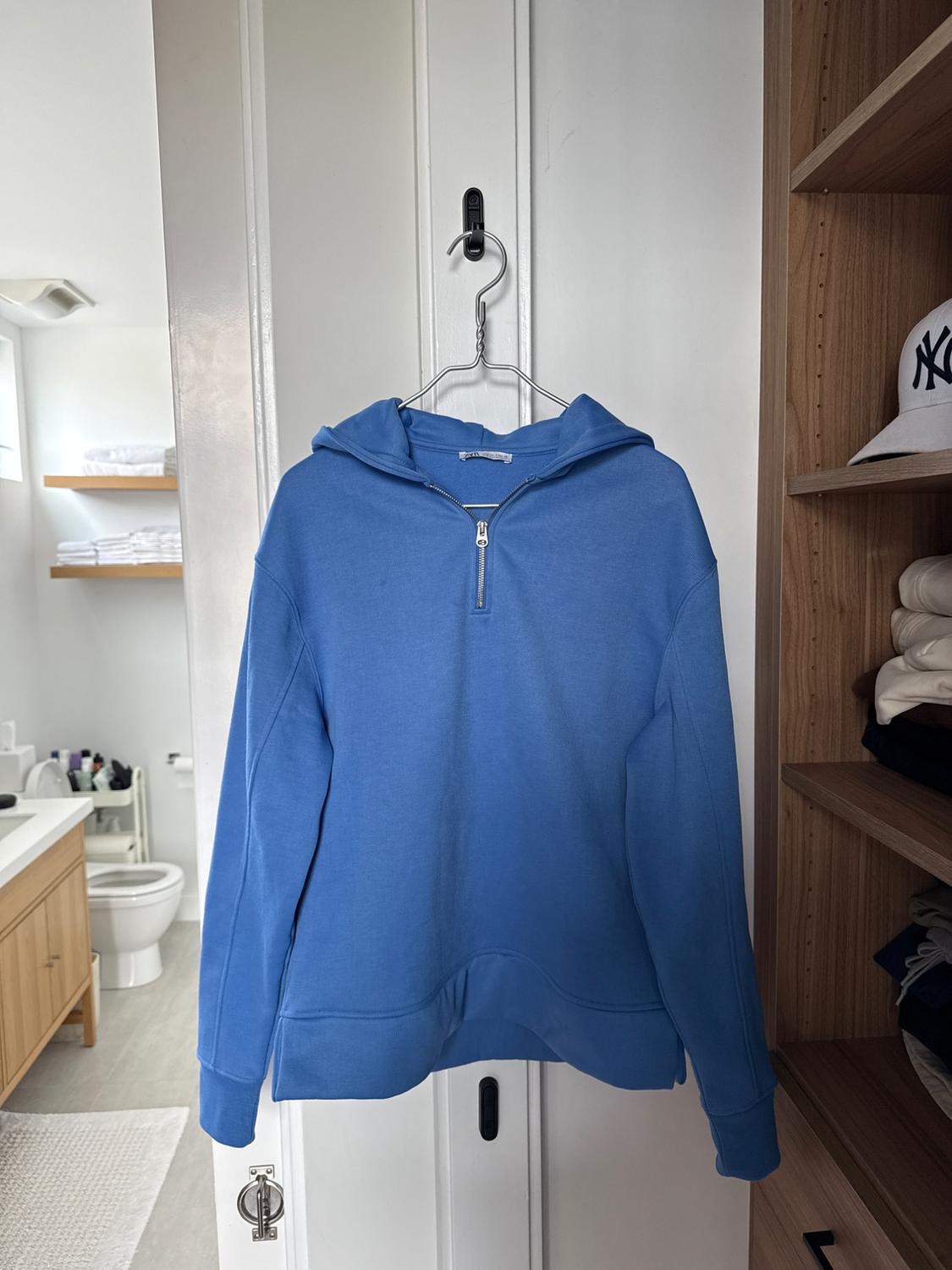 Blue zip-up hoodie hanging on a white door. Background with bathroom and wooden shelf with clothes and a white cap.