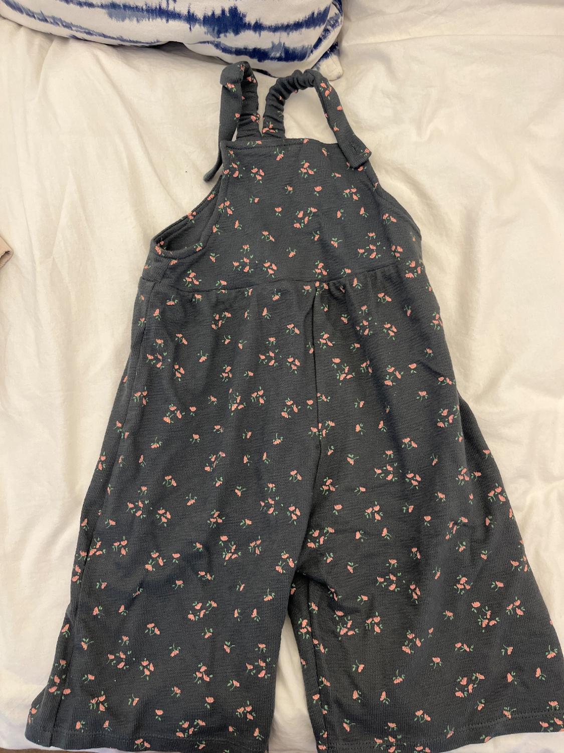 Dark grey jumpsuit with a pattern of small pink and green flowers, ruched straps, on a bed with white sheets and a blue cushion.