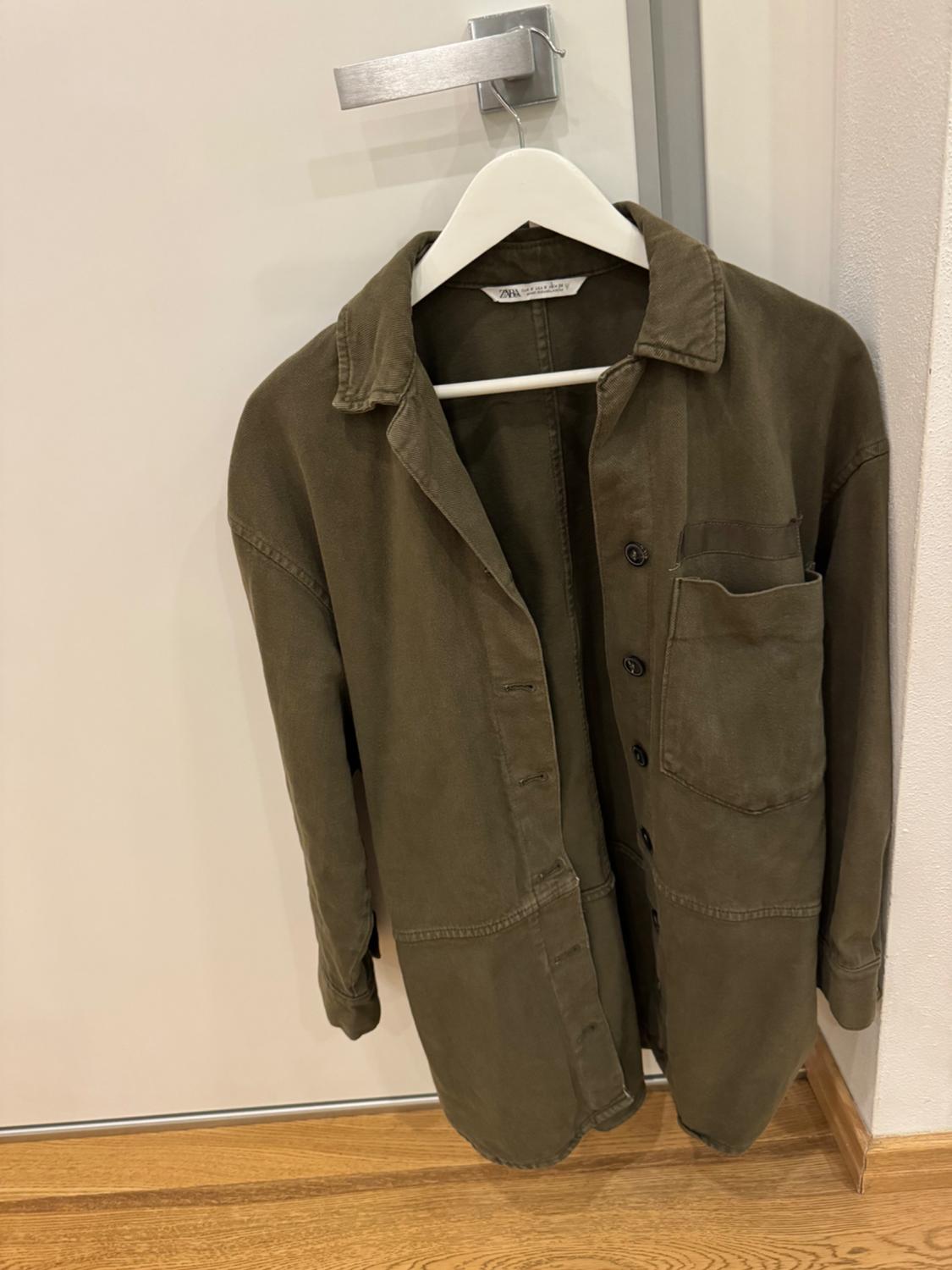 Olive green shirt hanging on a white hanger over a door. The label says "Zara Man". Wooden floor visible.