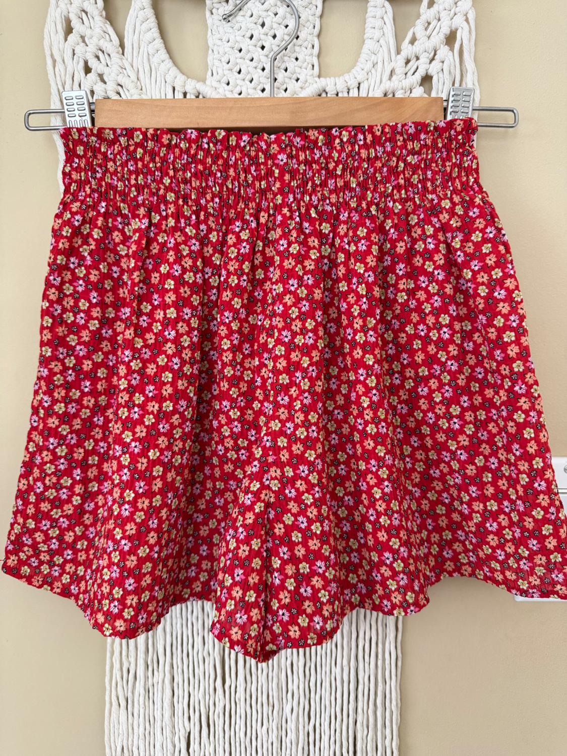 Red shorts with a small floral print in yellow and white tones, hanging on a hanger against a white macrame background.