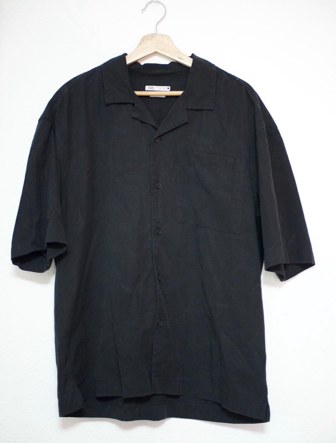 Short-sleeved black button-down shirt with a chest pocket, hanging on a wooden hanger against a white background. ZARA label.