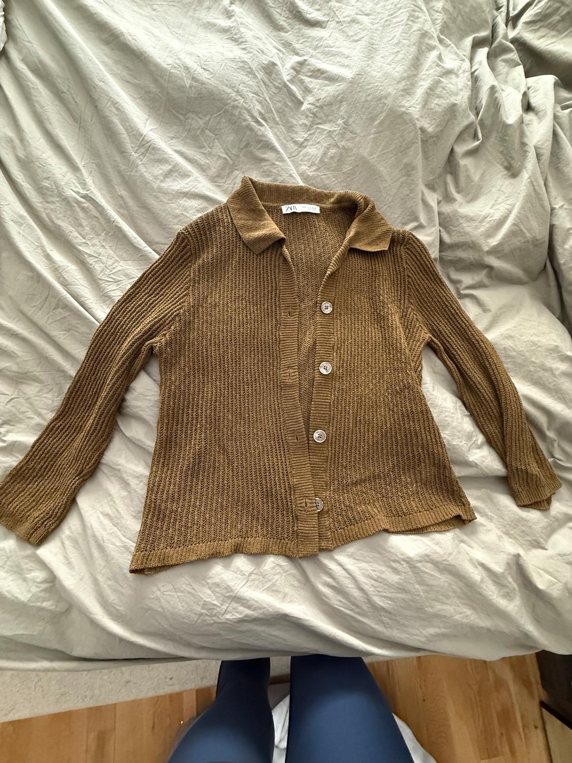 Brown knit cardigan with buttons, on a bed with wrinkled white sheets. Label visible on the collar.