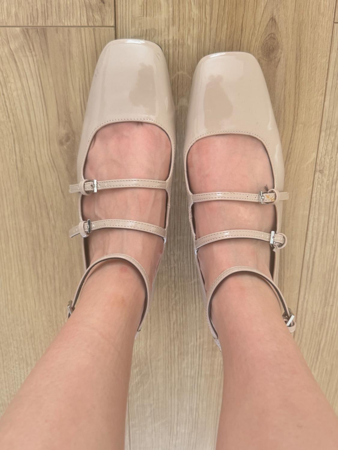 Beige patent leather shoes with a square toe and three straps, seen from above on a light wooden floor.
