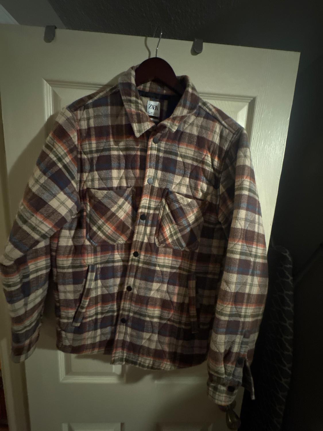 Tartan jacket in shades of brown, blue, and red hanging on a white door. Visible label with the text "ZARA".