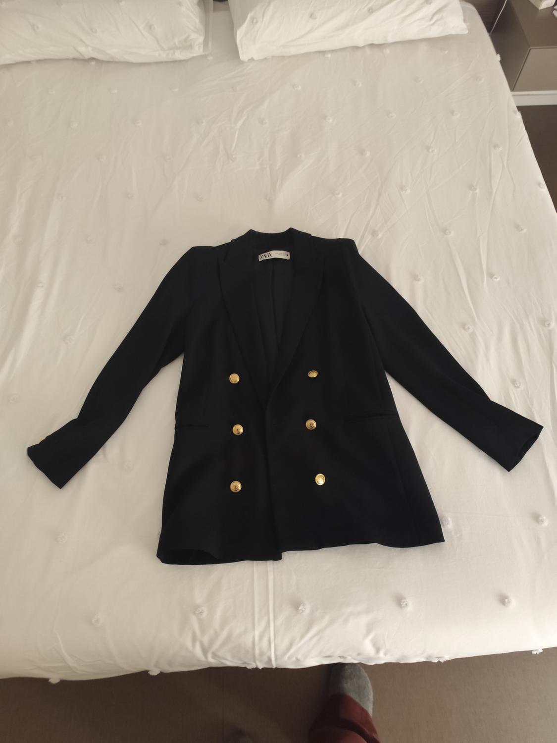 Black jacket with gold buttons on a white bed with pillows. Label visible on the jacket collar.