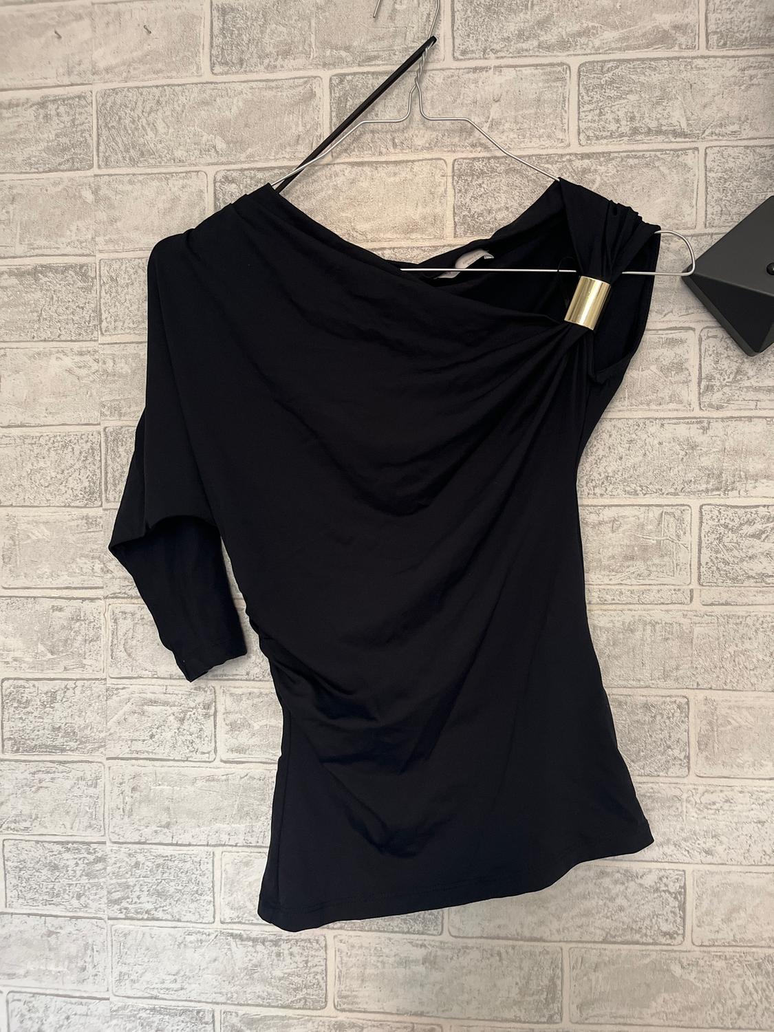 Black long-sleeved blouse hanging on a hanger against a white brick wall. Gold brooch detail on the shoulder.