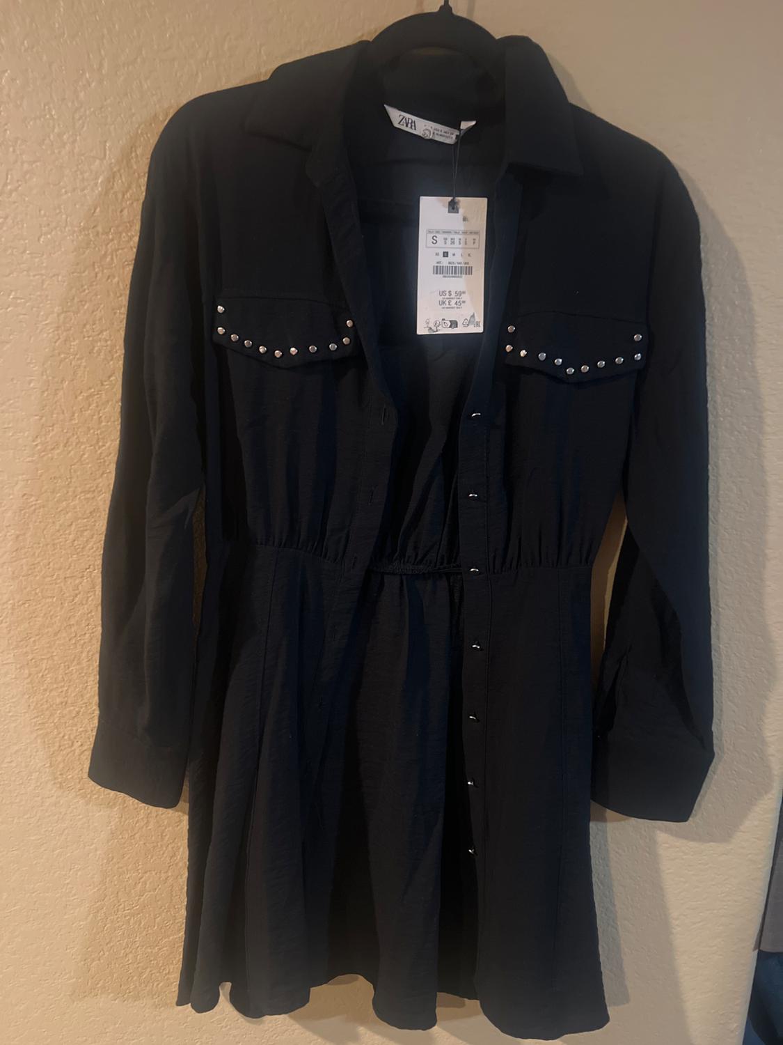 Black dress with buttons and studs on the chest pockets, hanging on a hanger. Label visible with price and size.