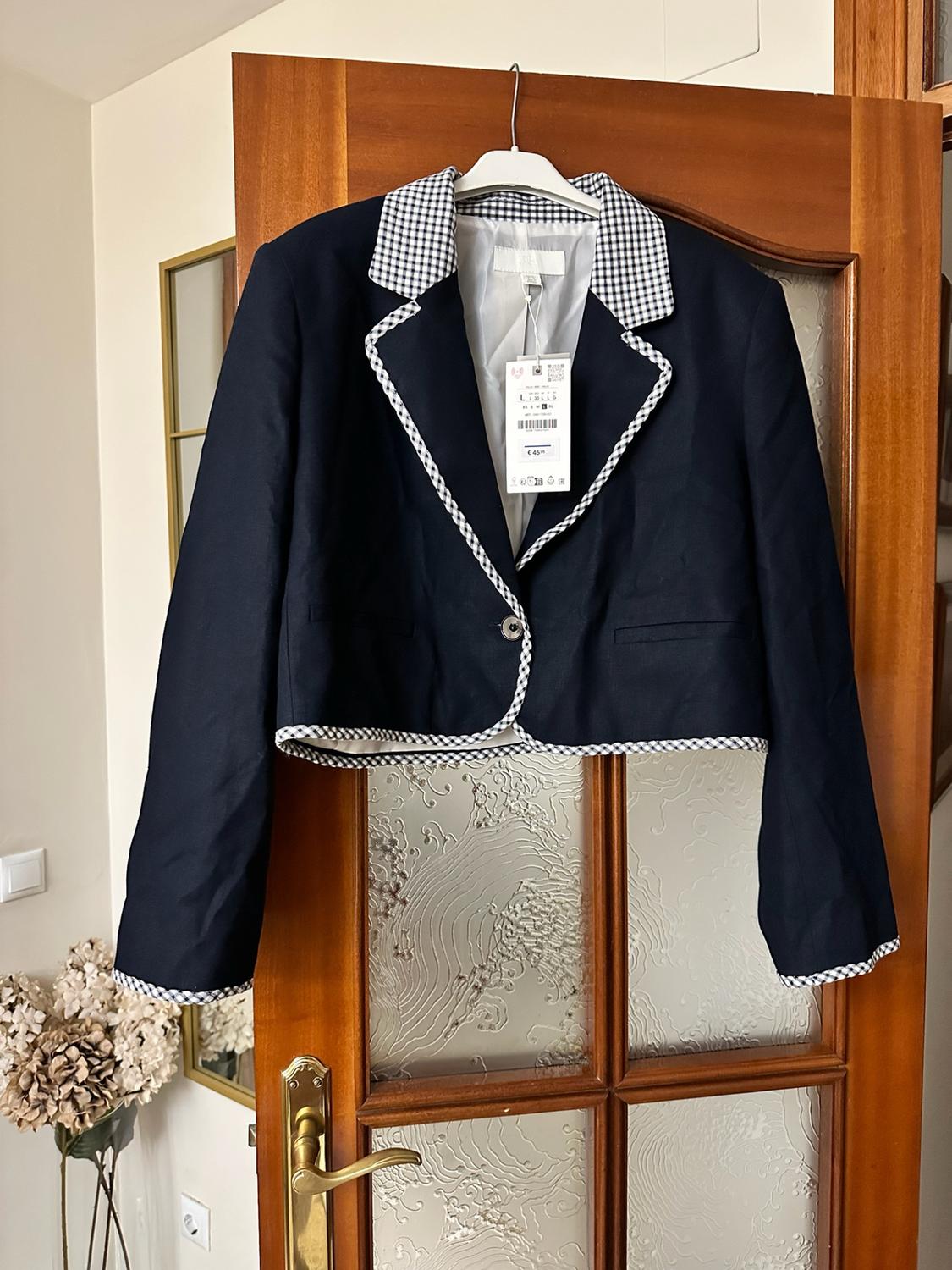 Short navy-blue jacket with a collar and black-and-white gingham trims, hanging on a wooden door.