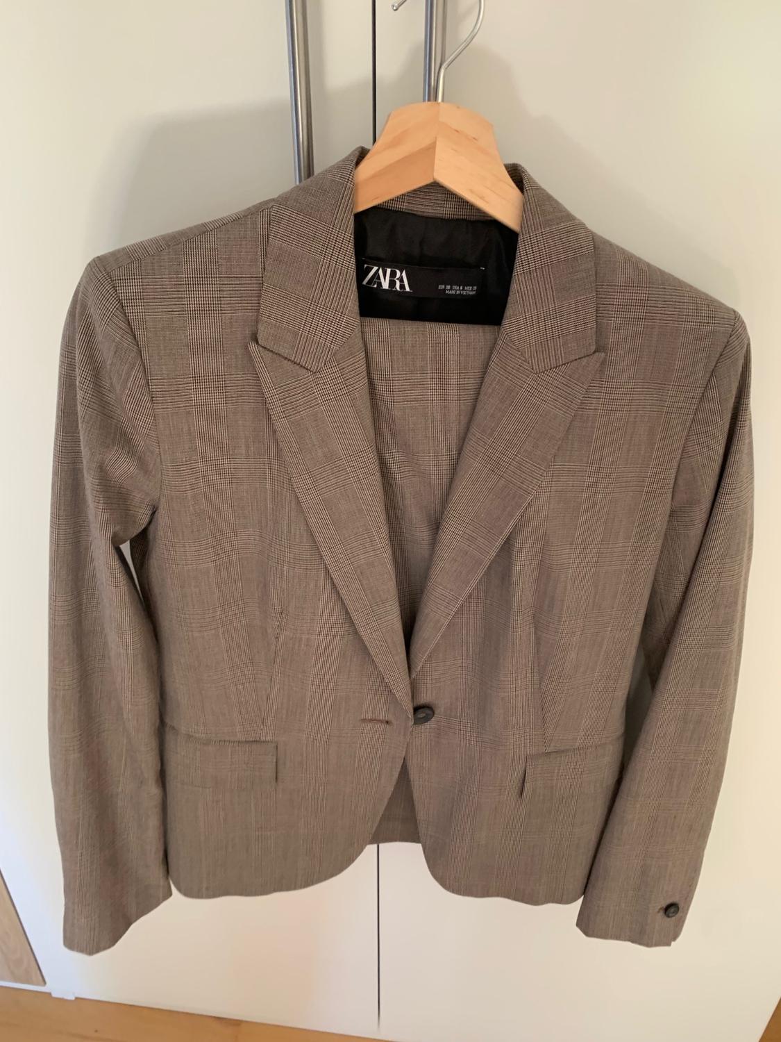 Checked blazer with lapels and pockets, hanging on a wooden hanger in front of a white wardrobe. Label with the text ZARA.
