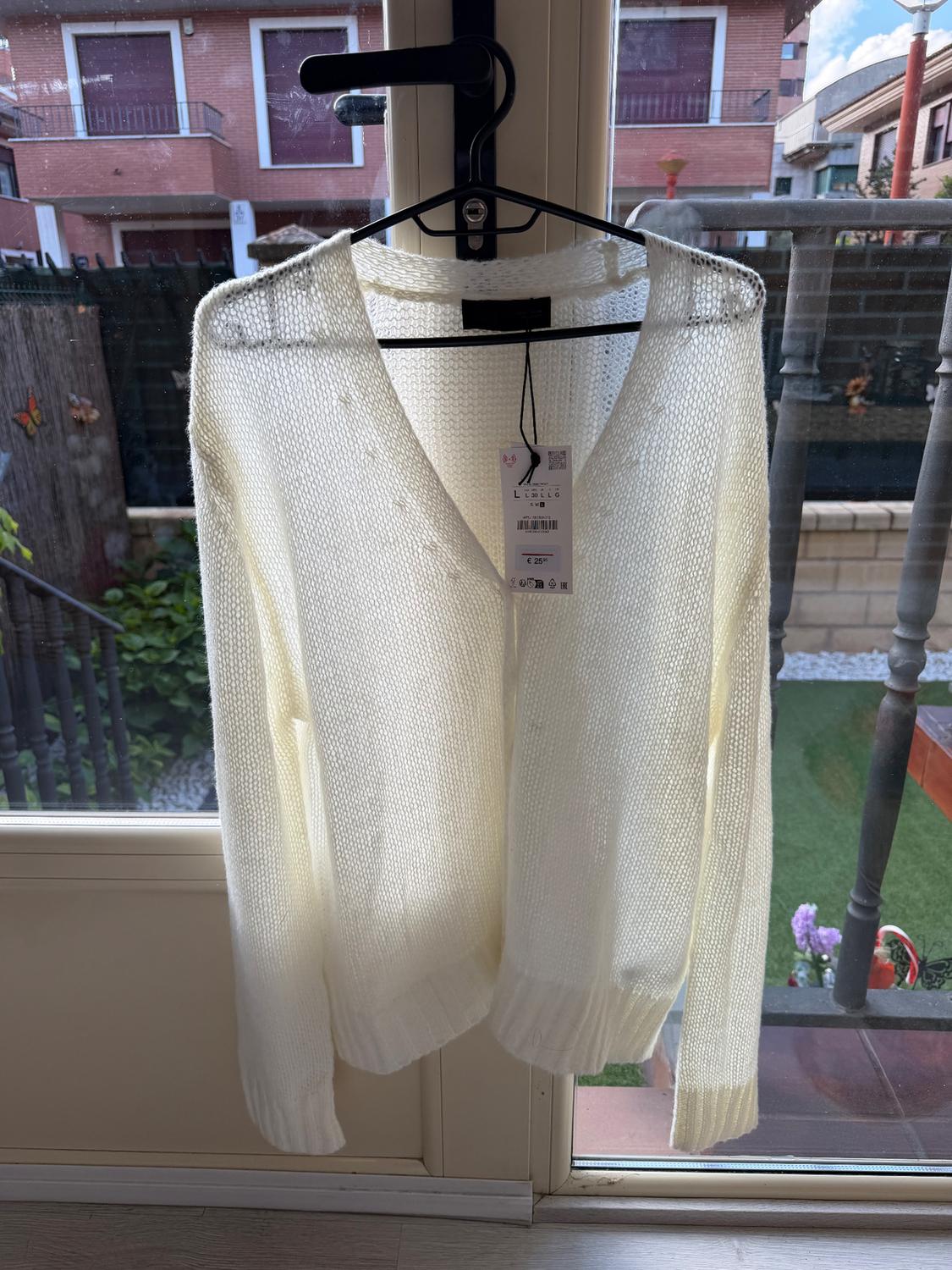 White knitted jumper hanging on a coat hanger in front of a window, with a visible label. Garden and building background.