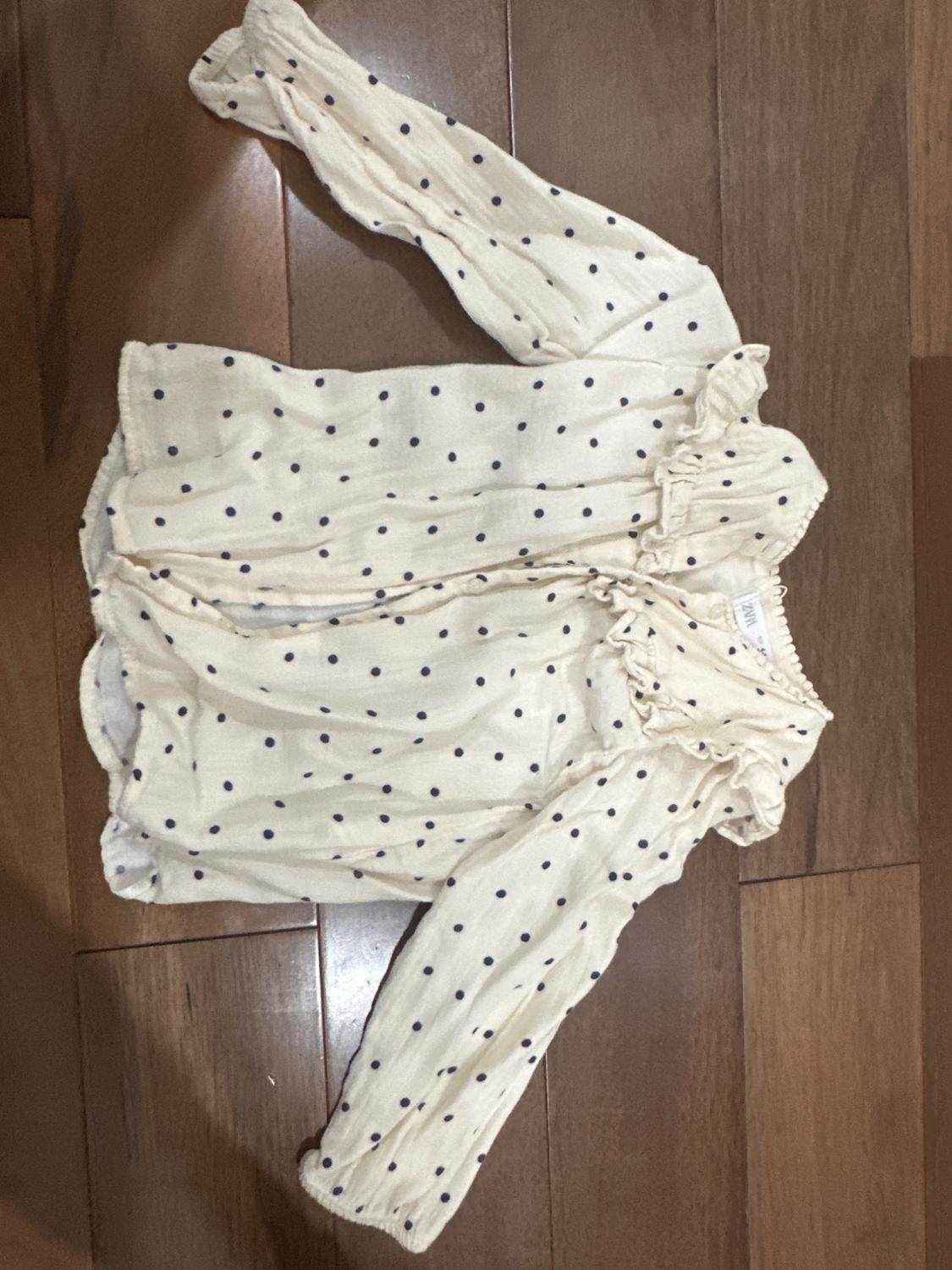 Cream-colored long-sleeved blouse with black polka dots on a wooden floor. Ruffle detail at the neck and cuffs.