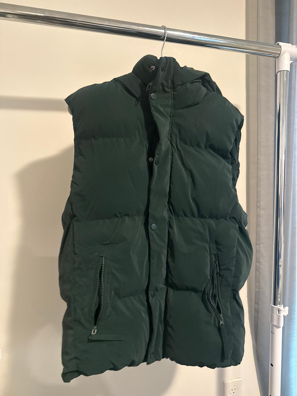 Dark green padded gilet hanging on a metal clothes rail. It has zipped pockets and press studs.