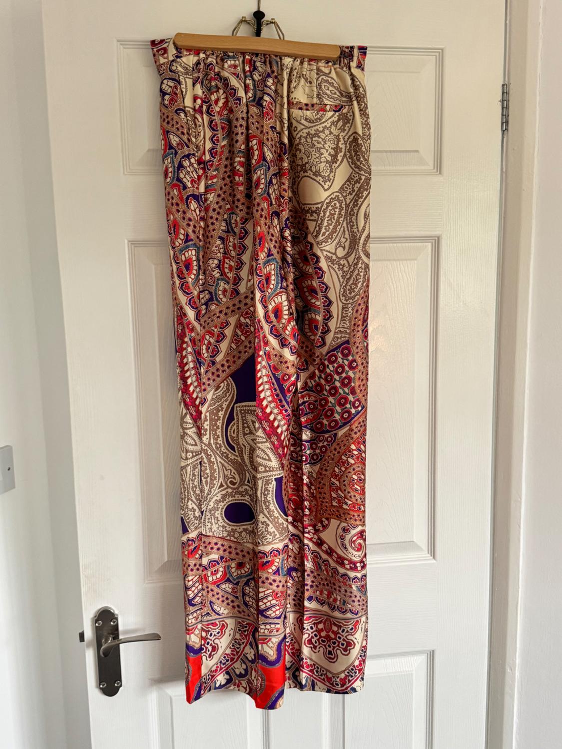Patterned trousers with a paisley design in beige, red, blue and purple tones, hanging on a hanger in front of a white door.
