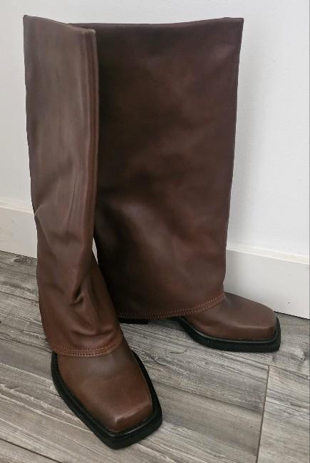 Brown square-toe high boots with a layered shaft, photographed on a grey wooden floor in front of a white wall.