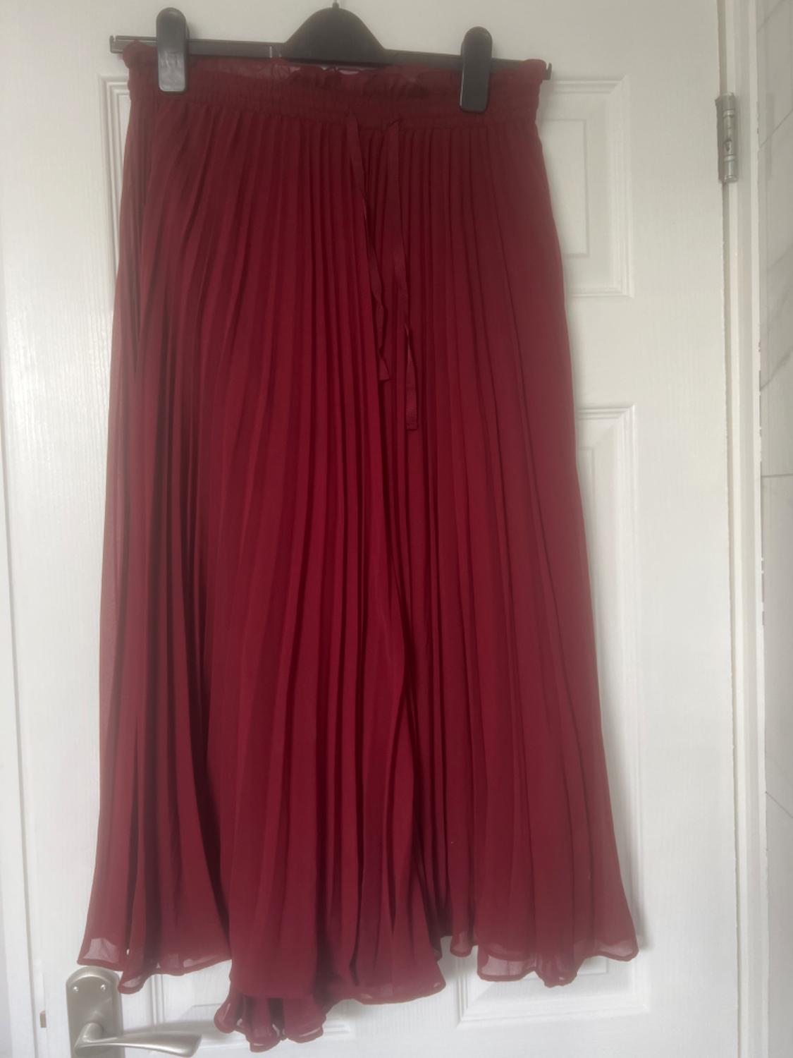 Red pleated skirt hanging on a white door. The fabric is light and flowing, with a tie at the waist.