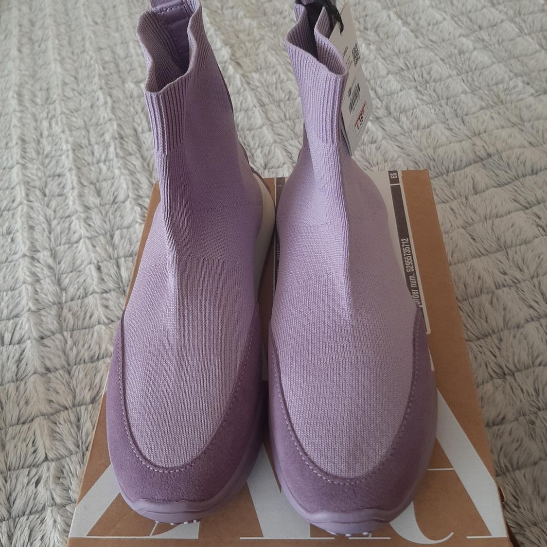 Ψηλά πλεκτά sneakers λιλά χρώματος πάνω σε ένα καφέ κουτί με ορατές ετικέτες, τοποθετημένα σε ένα γκρι χαλί.