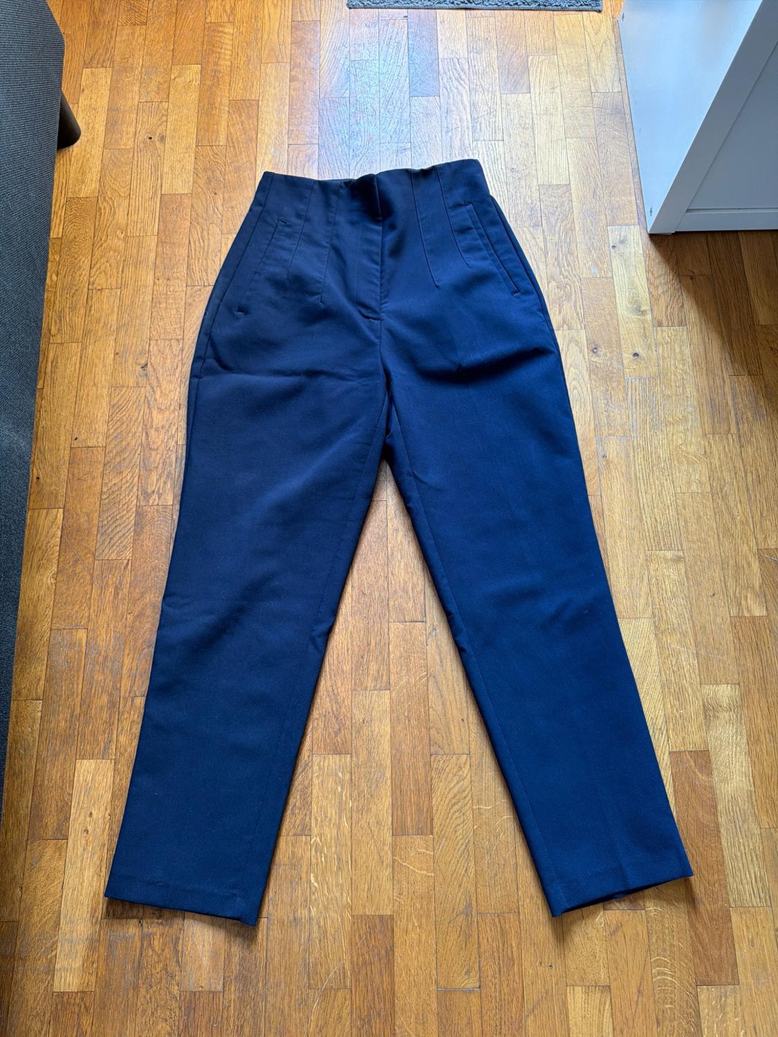 High-waisted navy-blue trousers with pleats and side pockets, laid out on a parquet floor.