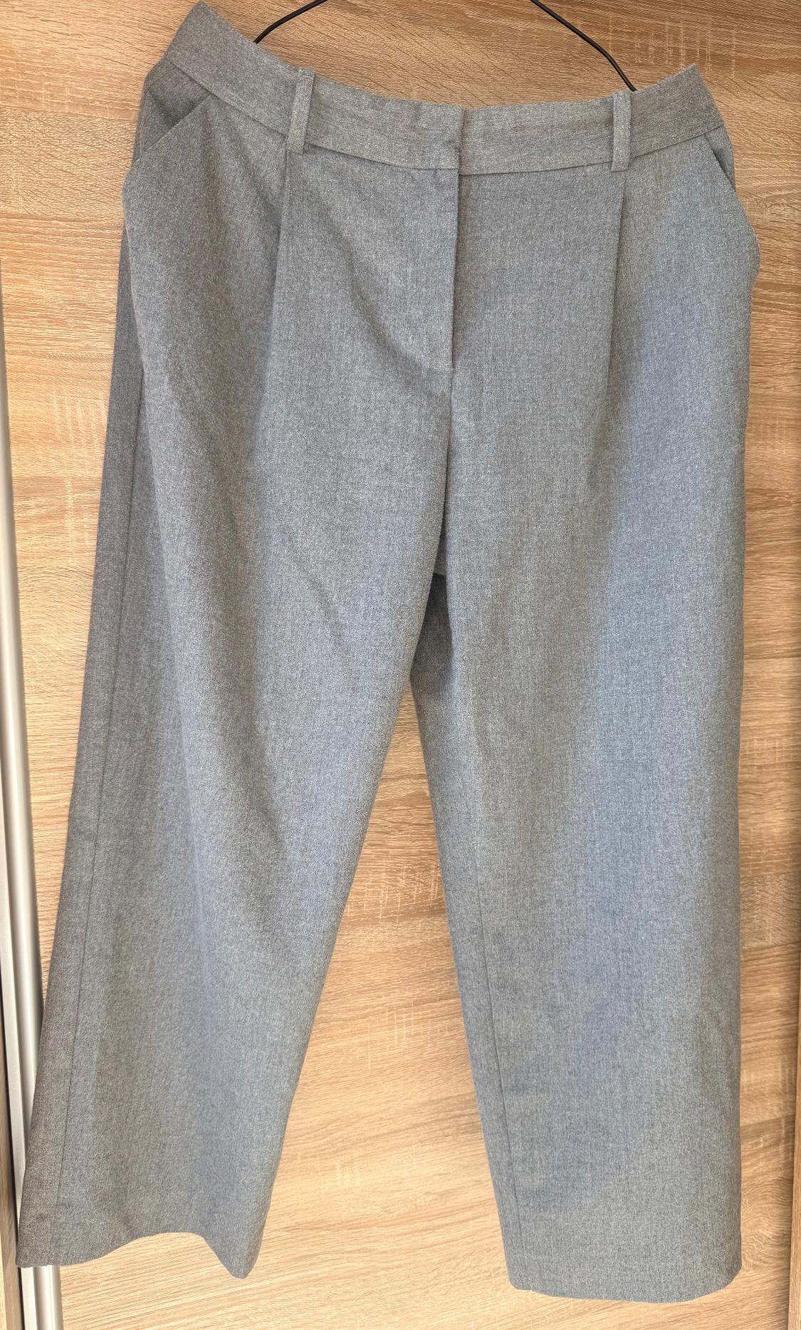 Grey fabric trousers hanging on a hanger against a light wooden background.