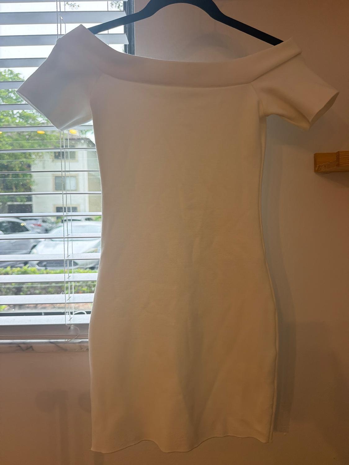 Short-sleeved, off-the-shoulder white mini dress, hanging on a black hanger next to a window with blinds.