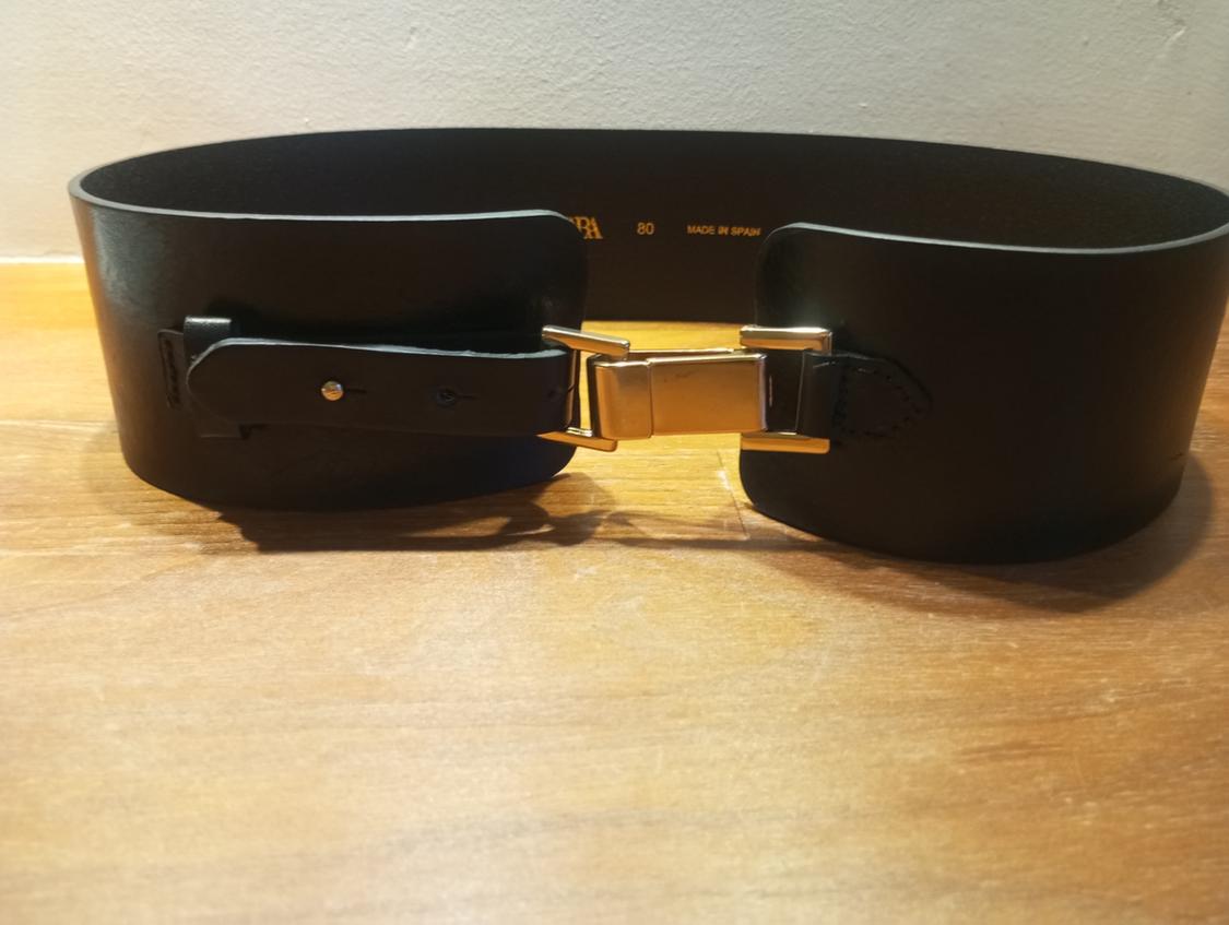 Wide black belt with a gold clasp on a wooden surface. Inside, the text "80 MADE IN SPAIN" is visible.