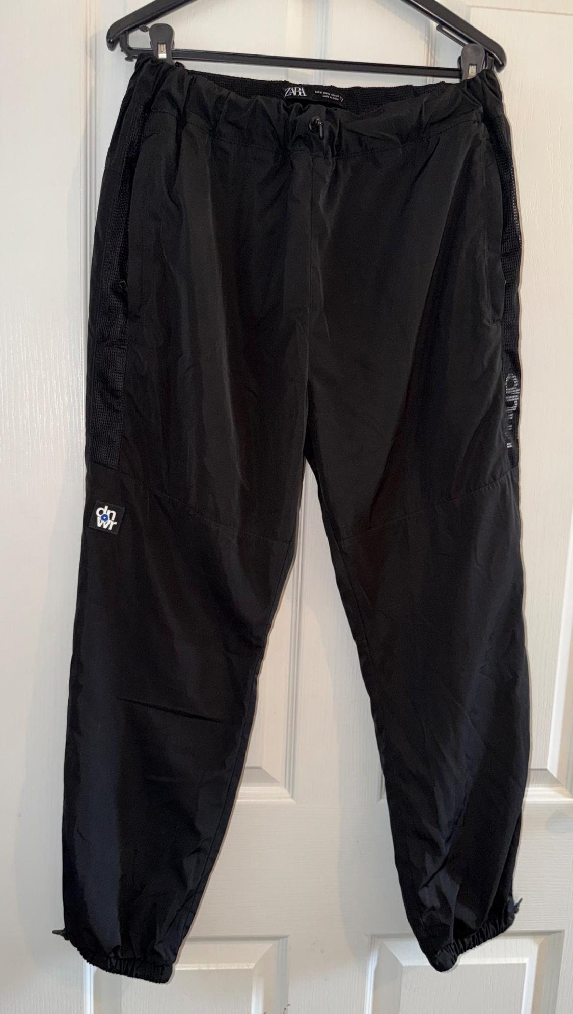 Black trousers hanging on a hanger, with elasticated waist and ankles. Visible label with 'dnvr' text on the left leg.