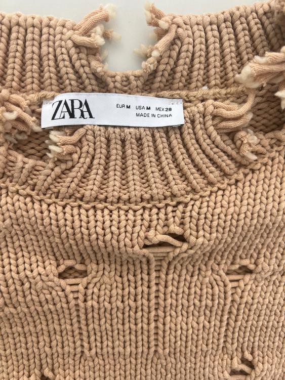 Zara tag on a beige knitted sweater with frayed details. Text: "EUR M USA M MEX 28 MADE IN CHINA".