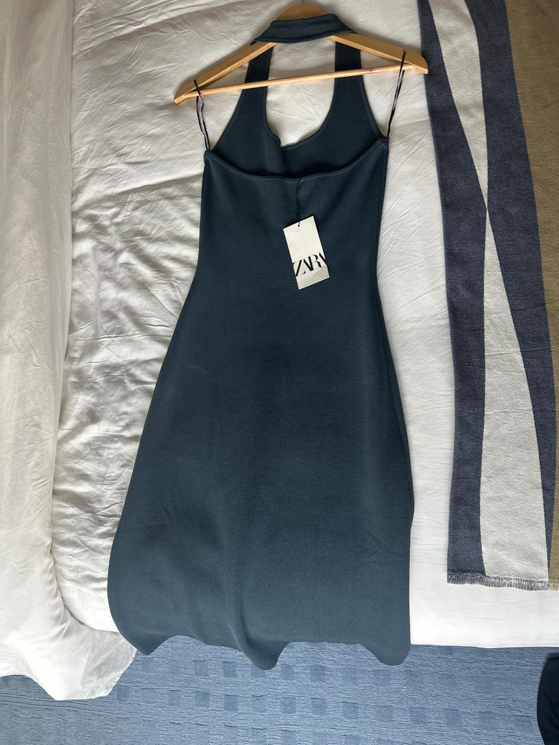 Sleeveless black dress on a hanger over a white bed, label with the text "ZARA". To the right, a blanket with blue and beige stripes.