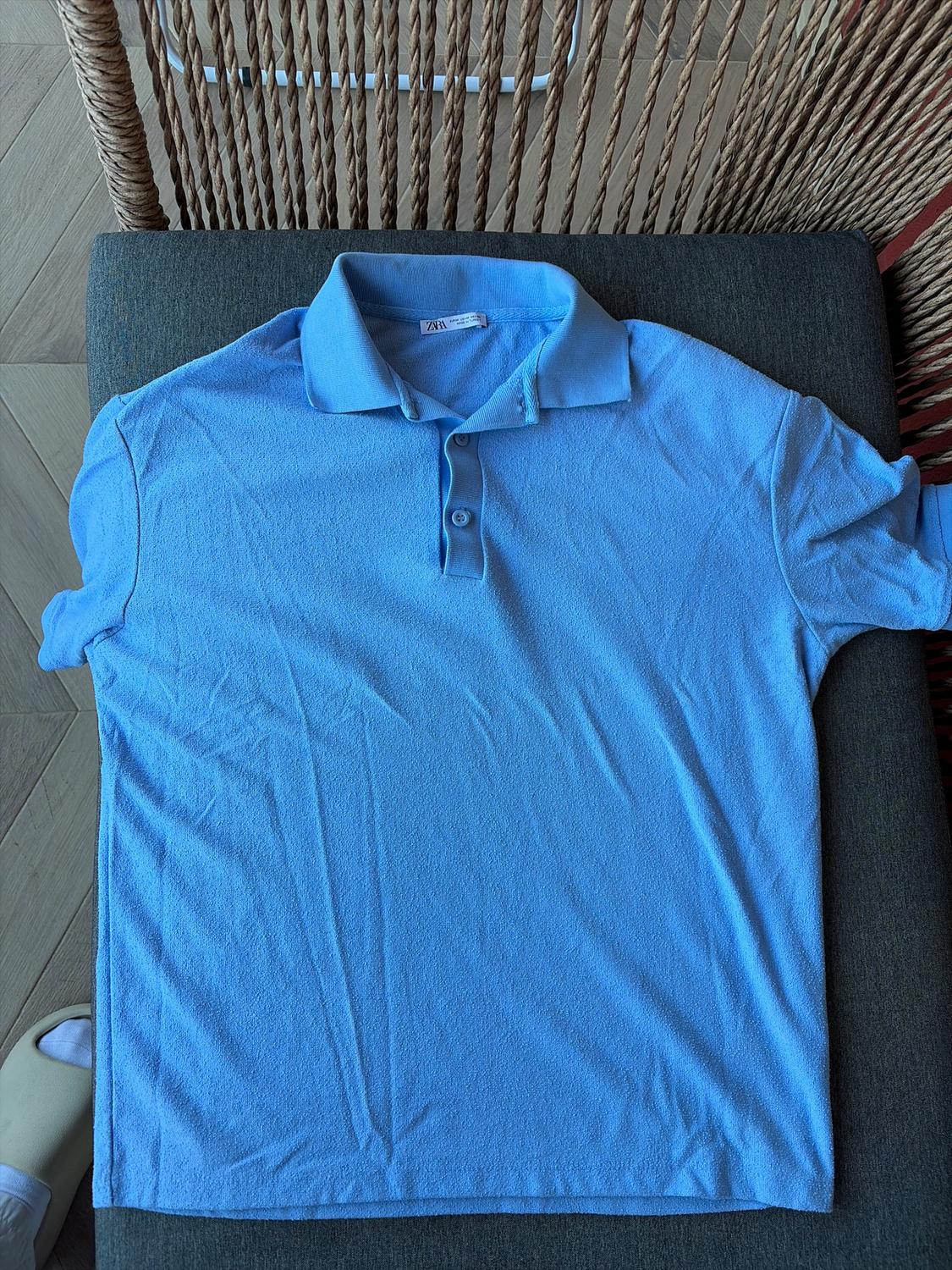 Light blue polo shirt with collar and buttons, placed on a grey cushion. Part of a rope chair visible in the background.