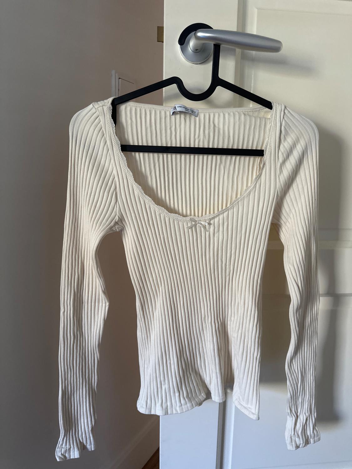 Top  ribbed μακρυμάνικο με στρογγυλή λαιμόκοψη, δαντέλα και φιόγκο μπροστά. Είναι κρεμασμένο σε μια κρεμάστρα στο πόμολο μιας λευκής πόρτας.