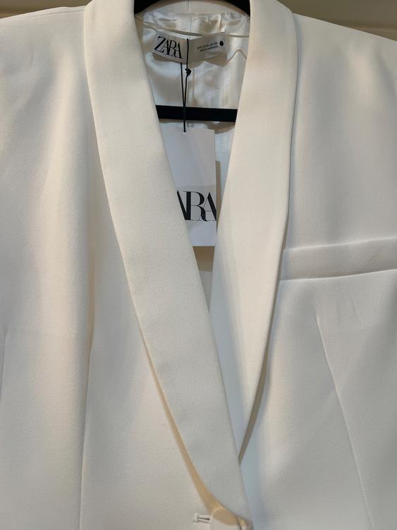 White suit jacket hanging on a hanger with a visible Zara tag. Soft fabric and elegant design.