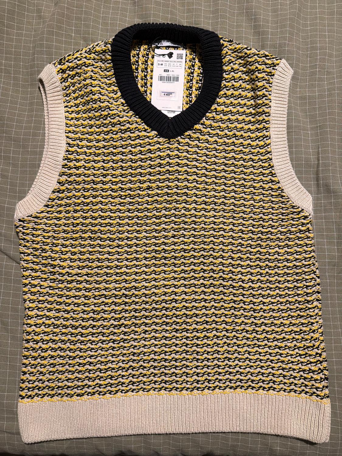 Sleeveless knitted vest with a woven pattern in yellow, black, and white. Black V-neck, visible label in the centre.