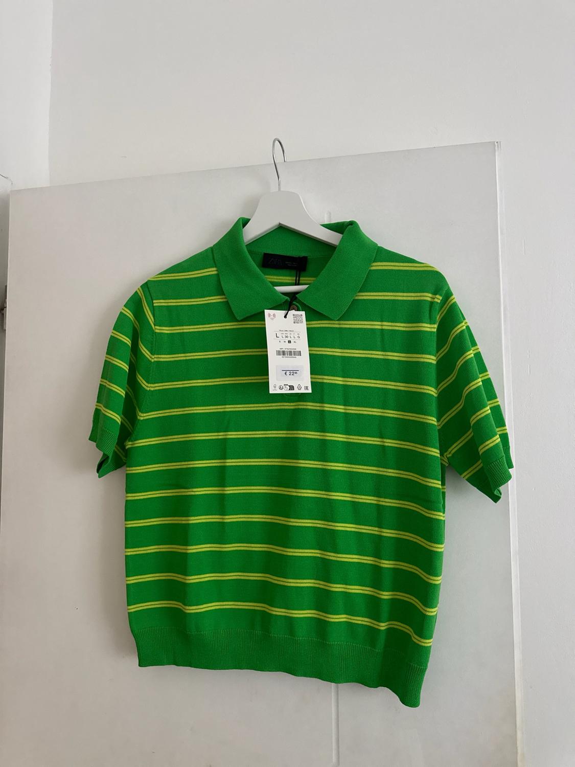 Green short-sleeved polo shirt with yellow stripes hanging on a white door. Label with text L and € 22.
