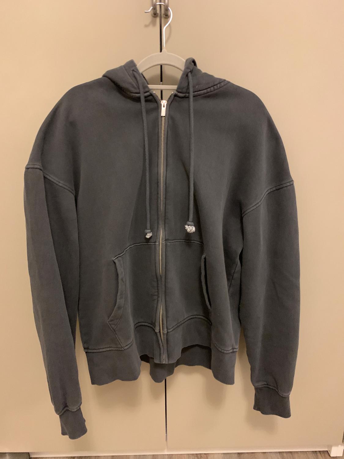 Grey hooded zip-up sweatshirt hanging on a hanger against a beige background. Front pockets and visible drawstrings.