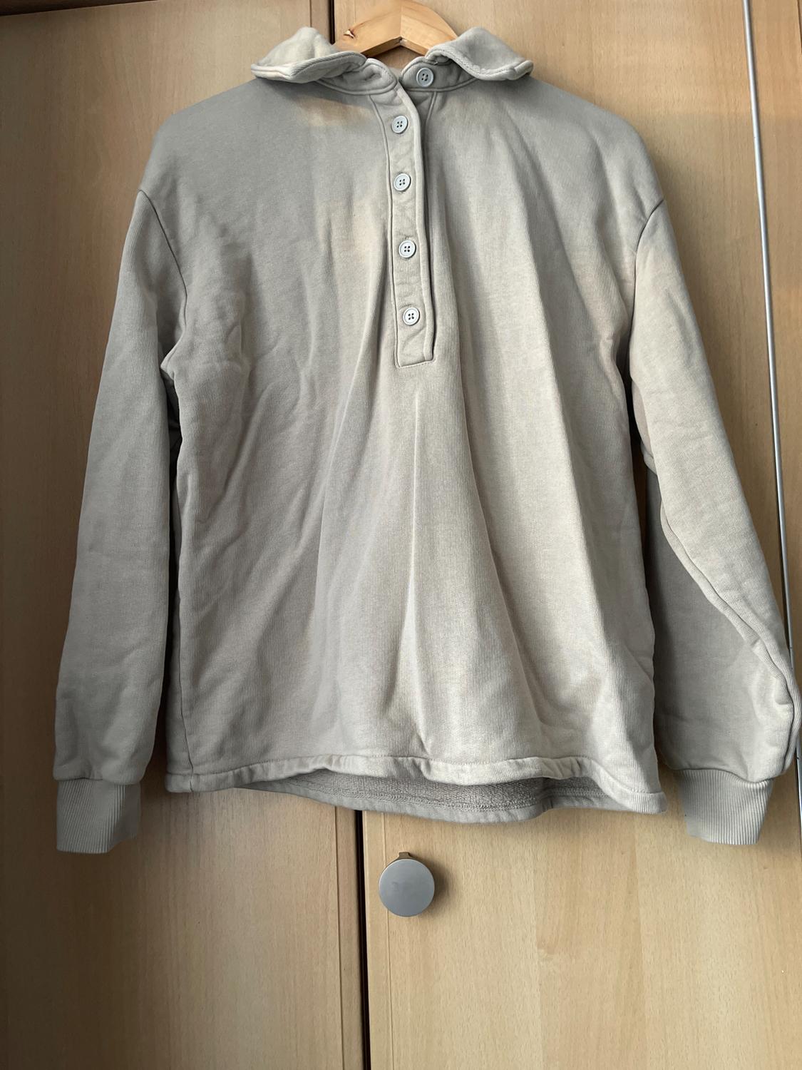 Beige long-sleeved sweatshirt with buttons on the neck, hanging on a wooden hanger in front of a light wood wardrobe.