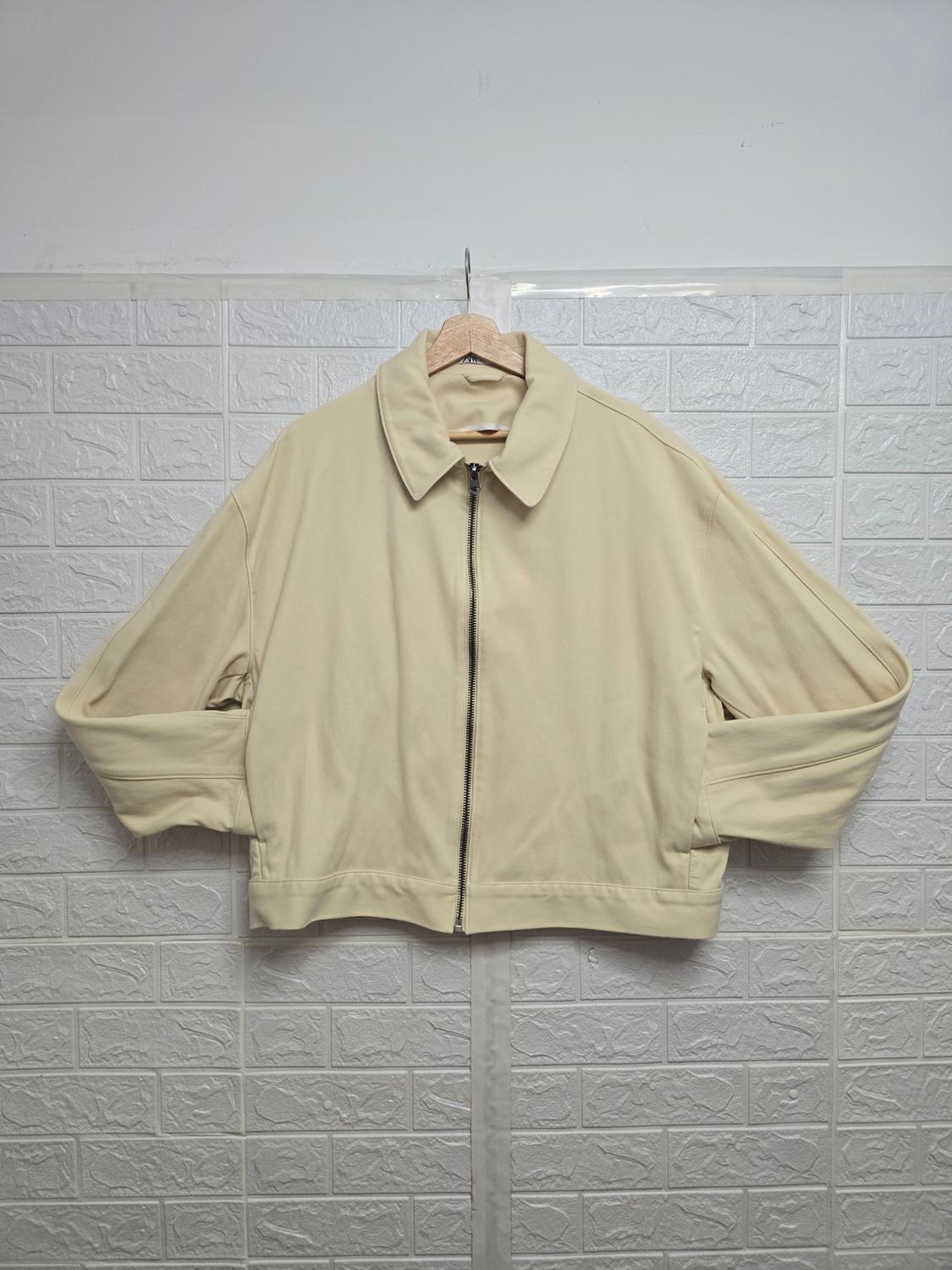 Beige jacket with a zip hanging on a wooden coat hanger, white brick-textured wall background.