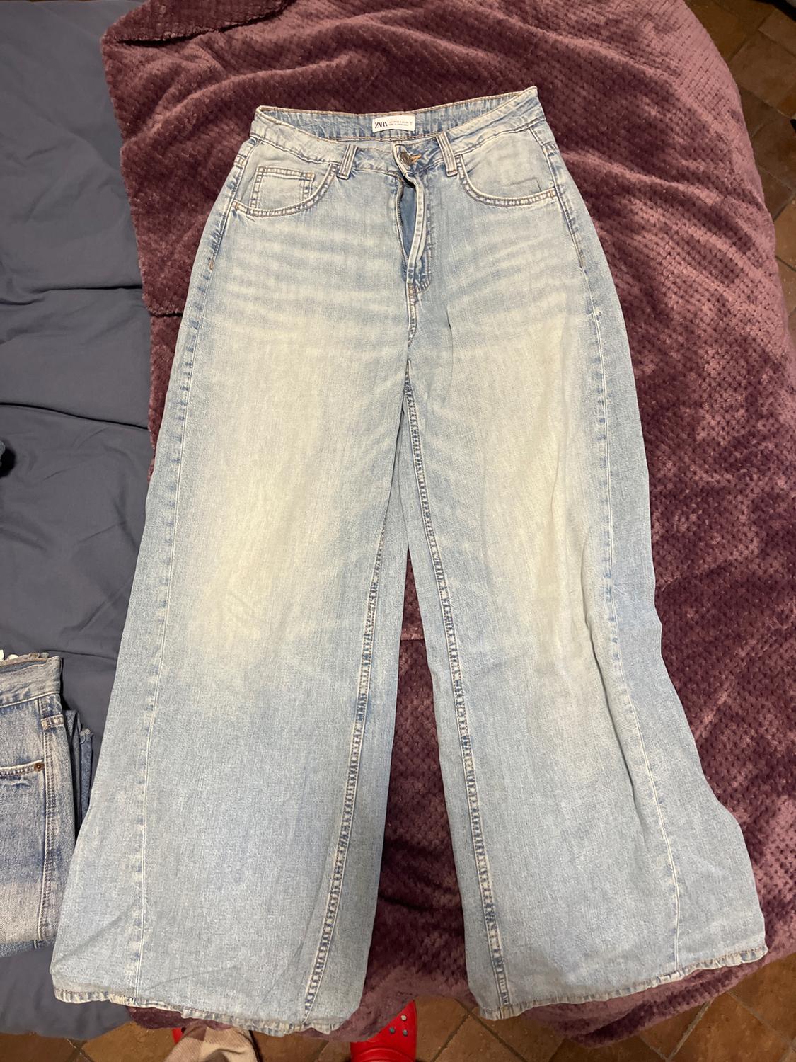 Light-wash denim wide-leg jeans on a purple blanket. Visible label on the waistband. Part of another pair of trousers on the left.